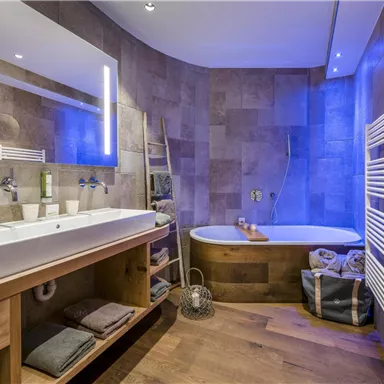 A modern bathroom with a double sink and a freestanding bathtub. The walls are clad with stone tiles, and the lighting creates a relaxing atmosphere.