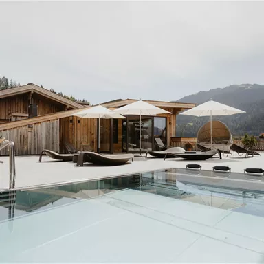 A modernly designed outdoor area with a pool, sun loungers, and umbrellas. In the background, beautiful mountains and wooden buildings can be seen.