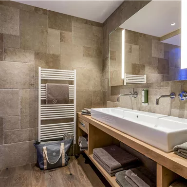 A modern bathroom with large, gray tiles and an elegant double sink. On the wall, there is a towel warmer and a wooden shelf with towels.