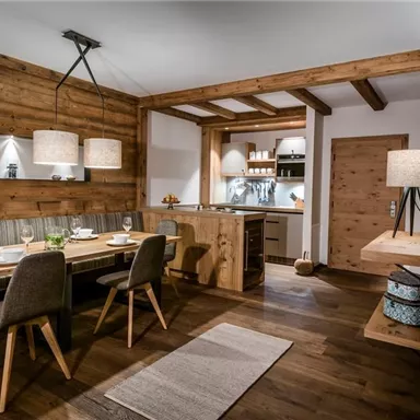 A modern dining room with wooden furniture and warm light accents. The open kitchen is well-equipped and invites to cook.