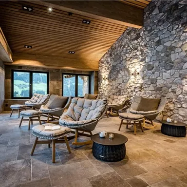 An elegant relaxation room with comfortable seating and large windows. The walls are made of stone, and the wooden ceiling creates a cozy atmosphere.