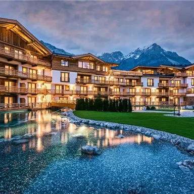 A modern hotel building with lit balconies. In the foreground, a tranquil pond surrounded by picturesque scenery and mountains in the background.