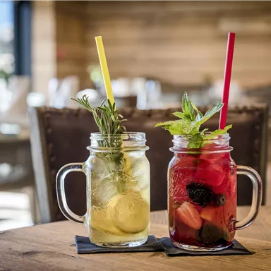 Two refreshing drinks in glasses on a wooden table. The left drink is light and decorated with lemon and herbs, while the right drink is colorful with berries and mint.