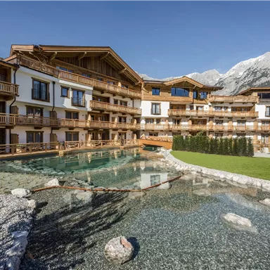 A modern wooden architecture in the mountains with a natural pond in the middle. Surrounded by green lawn and imposing mountain peaks.