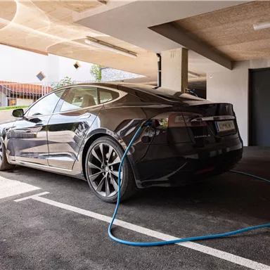 A black electric car is parked in a garage and is connected to a charging station. In the background, windows and a door are visible.