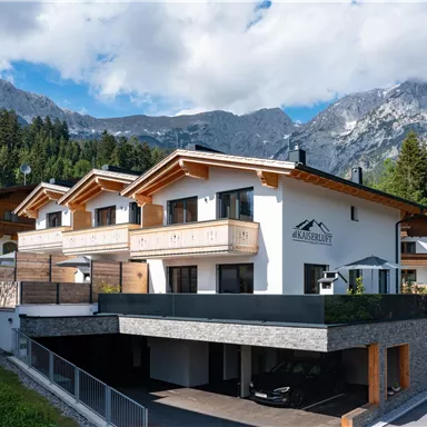 A modern house in the mountains with wooden siding and balconies. In the background, impressive mountains and a blue sky can be seen.