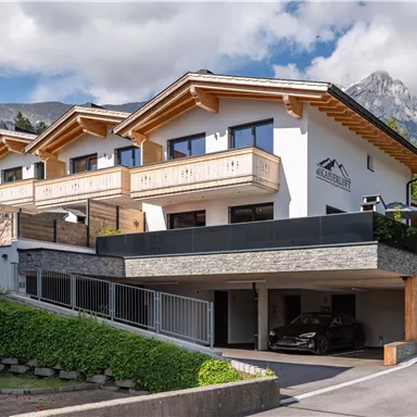 A modern building with wooden balustrades and a garage area. In the background, mountains and a green environment can be seen.