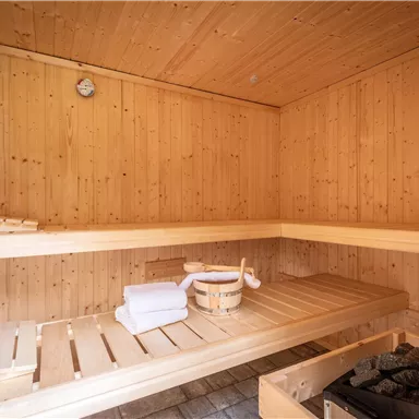 A cozy wooden sauna with benches and a small stove. Fresh towels and a wooden container are in the corner.