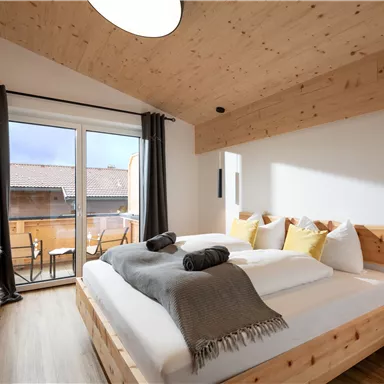 A cozy bedroom with wooden furniture and a large window. The decoration is simple and modern, with a balcony in the background.