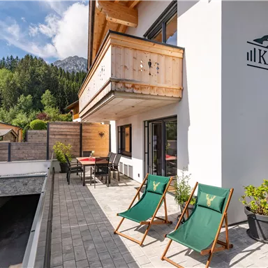 A modern chalet terrace with two lounge chairs and a view of a green landscape. The area is inviting and sunny, ideal for relaxing.