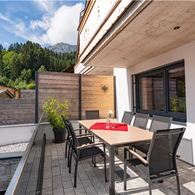 A modern terrace with a large table and chairs. Surrounded by green trees and a blue sky.