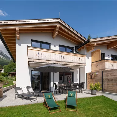 A modern mountain cabin with a spacious terrace and garden. In the background, majestic mountains and blue sky can be seen.