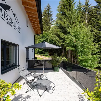 A modern terrace with loungers and a sun umbrella. Surrounded by lush greenery and trees, the place feels inviting and calm.