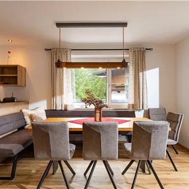 A modern dining room with a large table and gray chairs. Large windows allow plenty of daylight in and offer a view of the greenery.