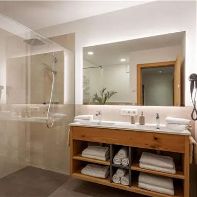 Ein modernes Badezimmer mit einer gläsernen Duschkabine und einem Holzwaschbecken. Auf dem Regal sind frische Handtücher und Pflegeprodukte ordentlich angeordnet.