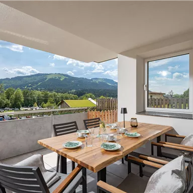 Eine gemütliche Terrasse mit einem Holztisch und Stühlen. Im Hintergrund sind grüne Berge und ein klarer Himmel zu sehen.