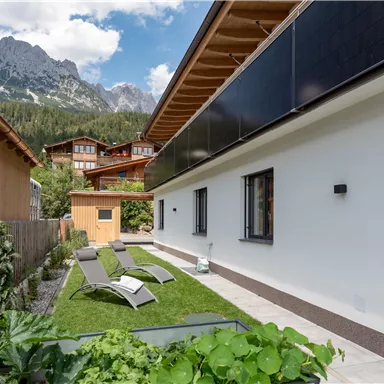 Ein ruhiger Garten mit Liegestühlen und einer modernen, weißen Wand. Im Hintergrund sind Berge und weitere Gebäude sichtbar.