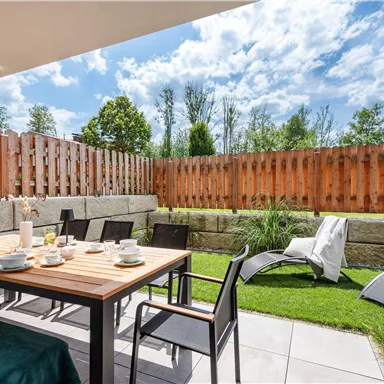 Eine gemütliche Terrasse mit einem Tisch, Stühlen und einem schönen Blick auf den Garten. Die Sonne scheint und der Himmel ist teilweise bewölkt.