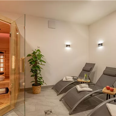 Ein entspannender Wellnessbereich mit Liegen und einer Sauna. Der Raum ist modern und hell gestaltet, mit einer Pflanze und frischen Äpfeln auf dem Tisch.