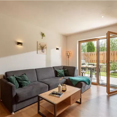 Ein modernes Wohnzimmer mit einem grauen Sofa und einem Holztisch. Große Fenster bieten einen Blick auf den Garten.