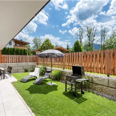 Ein gemütlicher Garten mit Rasen, einem Sonnenschirm und Liegen. Im Hintergrund ist eine Holzterrasse und ein Grill zu sehen.