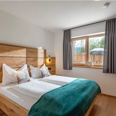 Ein gemütliches Schlafzimmer mit einem Doppelbett und modernen Holzmöbeln. Große Fenster sorgen für viel Tageslicht und einen Blick ins Freie.