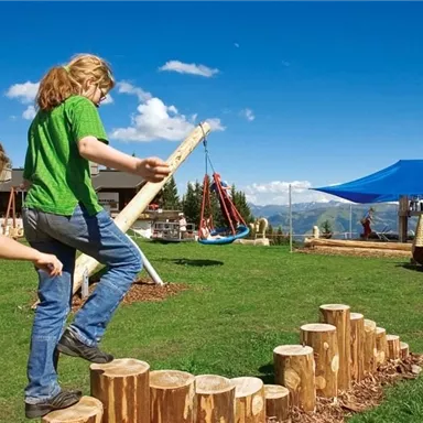Two children are playing outdoors on a playground with wooden logs. In the background, there are swings and a large blue tent.
