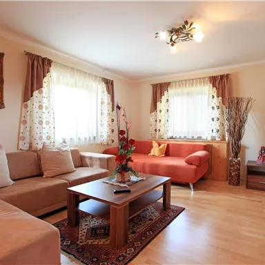 A bright living room with a beige sofa and a red sofa. Large windows let in a lot of light, and a table stands in the middle of the room.