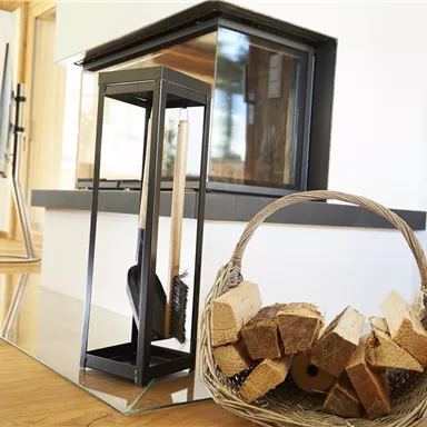 Ein moderner Kamin mit klaren Linien steht in einem hellen Raum. Neben dem Kamin befindet sich ein Korb mit Holzscheiten.