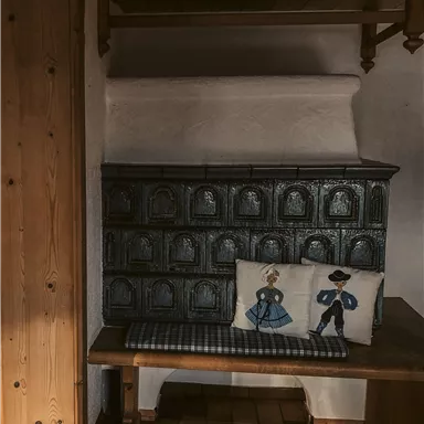 Eine gemütliche Ecke mit einer Holzbank und dekorativen Kissen. Im Hintergrund ist ein traditioneller Kachelofen zu sehen.