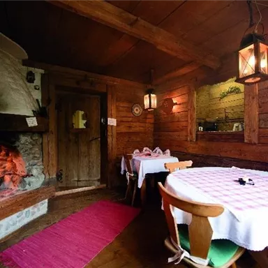 Eine gemütliche Stube mit Holzdekor und einem offenen Kamin. Zwei Tische sind mit einladenden Tischdecken gedeckt.