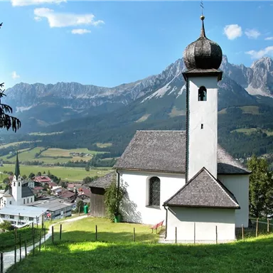 A picturesque church on a hill with an impressive mountain panorama in the background. In the foreground, you can see a green meadow and the village in the valley.