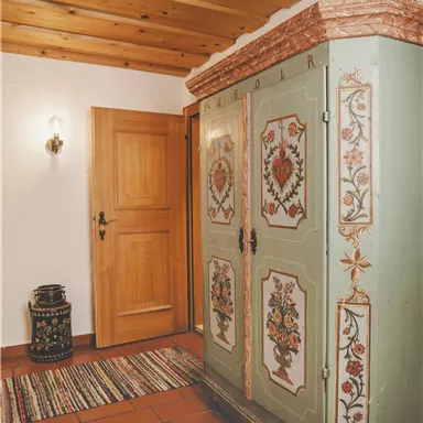 Ein dekorativer Kleiderschrank mit bunten Blumenmustern steht in einem Raum. Im Hintergrund sieht man eine Holztür und einen Teppich auf dem Boden.