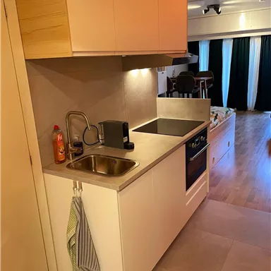 A modern kitchen with a sink, a stove, and storage space. The room is brightly lit and has a minimalist style.