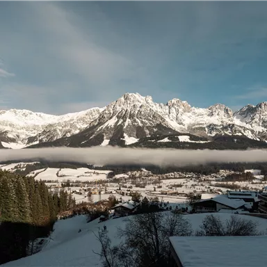 Eine beeindruckende Mountainlandschaft mit schneebedeckten Gipfeln und sanften Wolken. Im Vordergrund sind schneebedeckte Hügel und eine bewaldete Fläche zu sehen.
