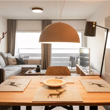 Ein modernes Esszimmer mit einem Holztisch und stilvollen Platzdeckchen. Im Hintergrund befindet sich ein gemütlicher Wohnbereich mit einem grauen Sofa und großen Fenstern.