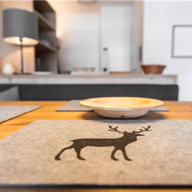 Ein eleganter Esstisch mit Tischsets aus Filz, die ein Reh-Motiv zeigen. Im Hintergrund sind sanfte Möbel und eine gemütliche Raumbeleuchtung zu sehen.