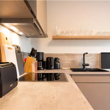 Eine moderne Küche mit hellen Farben und klaren Linien. Auf der Arbeitsfläche stehen Kochutensilien und Haushaltsgeräte.