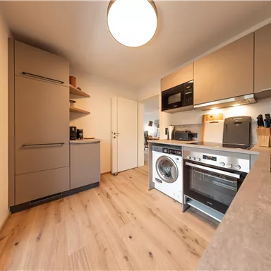 Eine moderne Küche mit hellen Schränken und weißen Geräten. Der Fußboden ist aus Holz und das Gesamtbild ist freundlich und einladend.