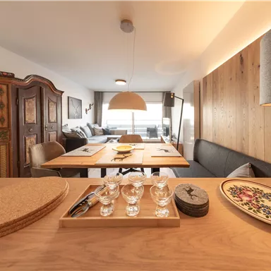 Ein gemütlicher Essbereich mit einem Holztisch und eleganten Geschirr. Im Hintergrund ist ein modernes Wohnobjekt mit einer großen Fensterfront zu sehen.