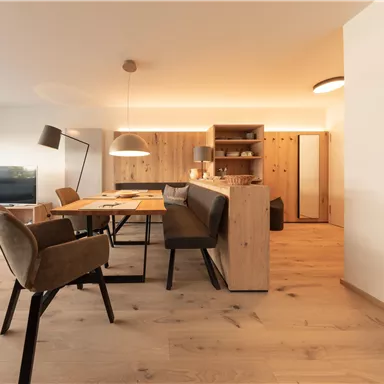 Ein modernes Wohnzimmer mit einem Esstisch und komfortablen Stühlen. Der Raum ist hell und freundlich, mit Holzelementen und großen Fenstern.