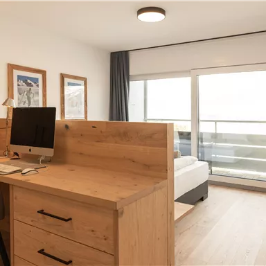Ein modernes Zimmer mit Holzdetails und einem Schreibtisch. Bodentiefe Fenster bieten einen Blick auf einen Balkon.
