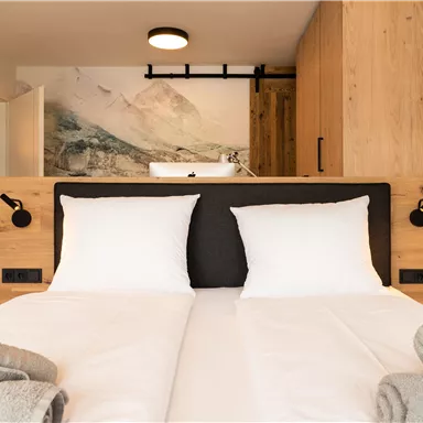 Ein modernes Schlafzimmer mit einem Doppelbett und weißen Kissen. Die Wände sind in warmen Holzfarben gehalten, und es gibt eine malerische Wandgestaltung im Hintergrund.