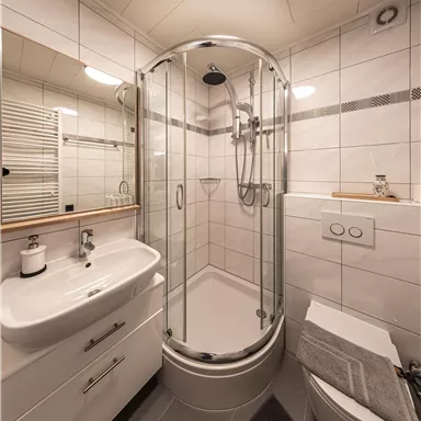 Ein modernes Badezimmer mit einer gläsernen Dusche und einem Waschbecken. Die Wände sind mit weißen Fliesen verkleidet und es ist gut beleuchtet.