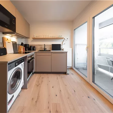 Eine moderne Küche mit einheitlichen Möbeln und einer Waschmaschine. Große Fenster bieten Blick auf einen Balkon mit Tisch und Stühlen.