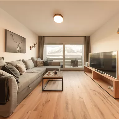 Ein modernes Wohnzimmer mit einem bequemen Sofa und einem großen Fernseher. Der Raum hat eine helle, einladende Atmosphäre und große Fenster mit Blick auf die Berge.