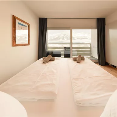 Ein modernes Hotelzimmer mit zwei getrennten Betten und großen Fenstern. Der Blick zeigt schneebedeckte Berge im Hintergrund.