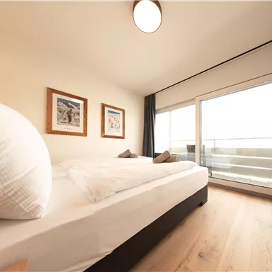 Ein gemütliches Schlafzimmer mit einem großen Bett und einem Fenster, das auf einen Balkon führt. Die Wände sind mit Bildern dekoriert, und das Zimmer ist hell und einladend.