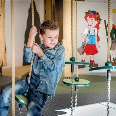 A boy is sitting on a swing and smiling. In the background, childlike figures can be seen on the wall.