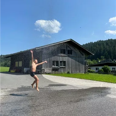 Ein Junge springt unter einem Wasserstrahl vor einem Holzhaus. Der Himmel ist blau und die Umgebung ist grün.